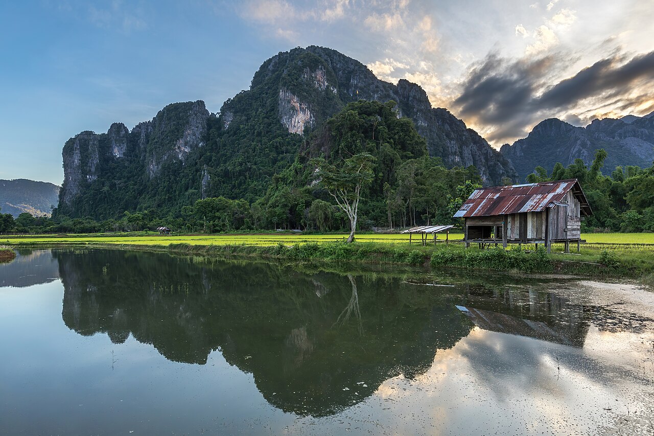 Water reflection of karst mountains, wooden hut, trees and colorful clouds at sunset with green paddy fields, Vang Vieng, Laos, illustrative activity photo